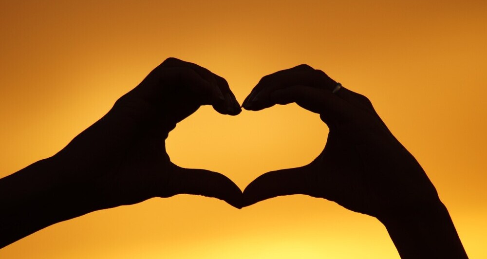 two hands making a heart in front of a yellow sky