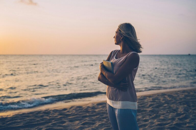 Woman standing on a beach looking at the sunset holding a journal. she looks pleased