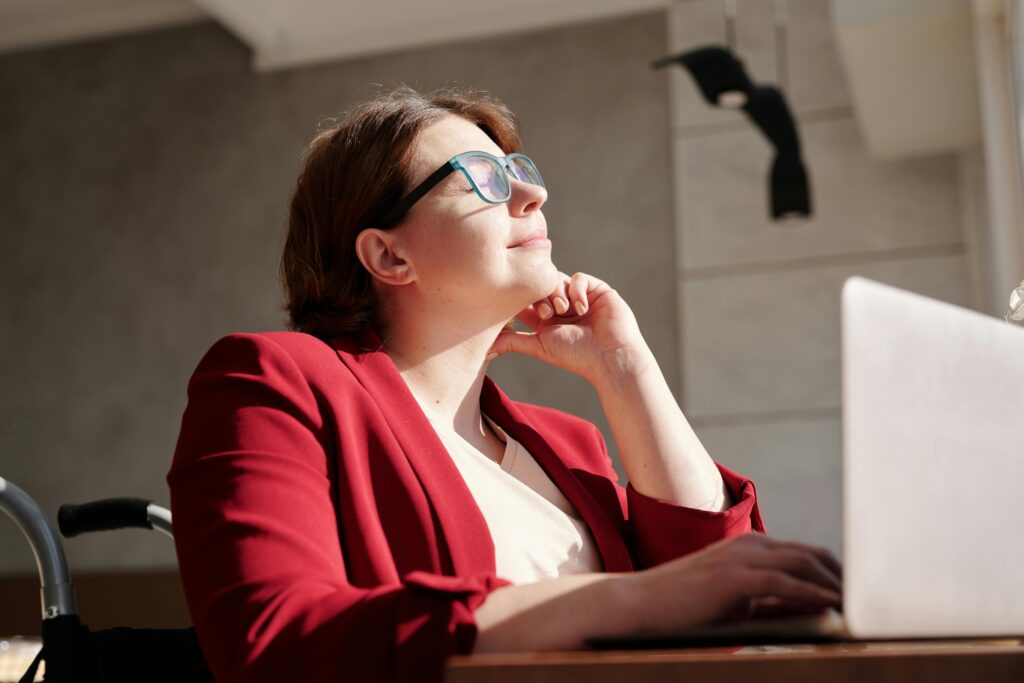 Woman in a wheelchair sitting at a desk with a laptop in front of her. she is basking in the sunlight