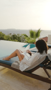 A woman sits by the pool on a chair reading a book