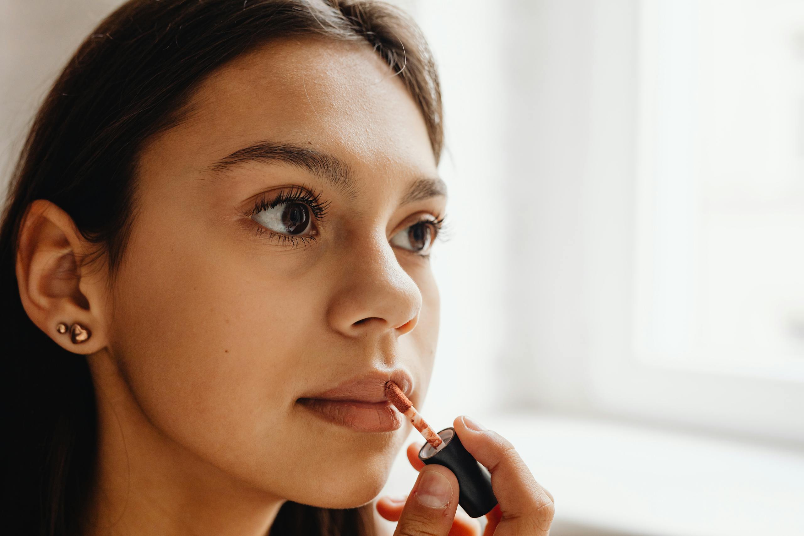 Close-up of a young woman applying lipstick indoors with natural daylight.