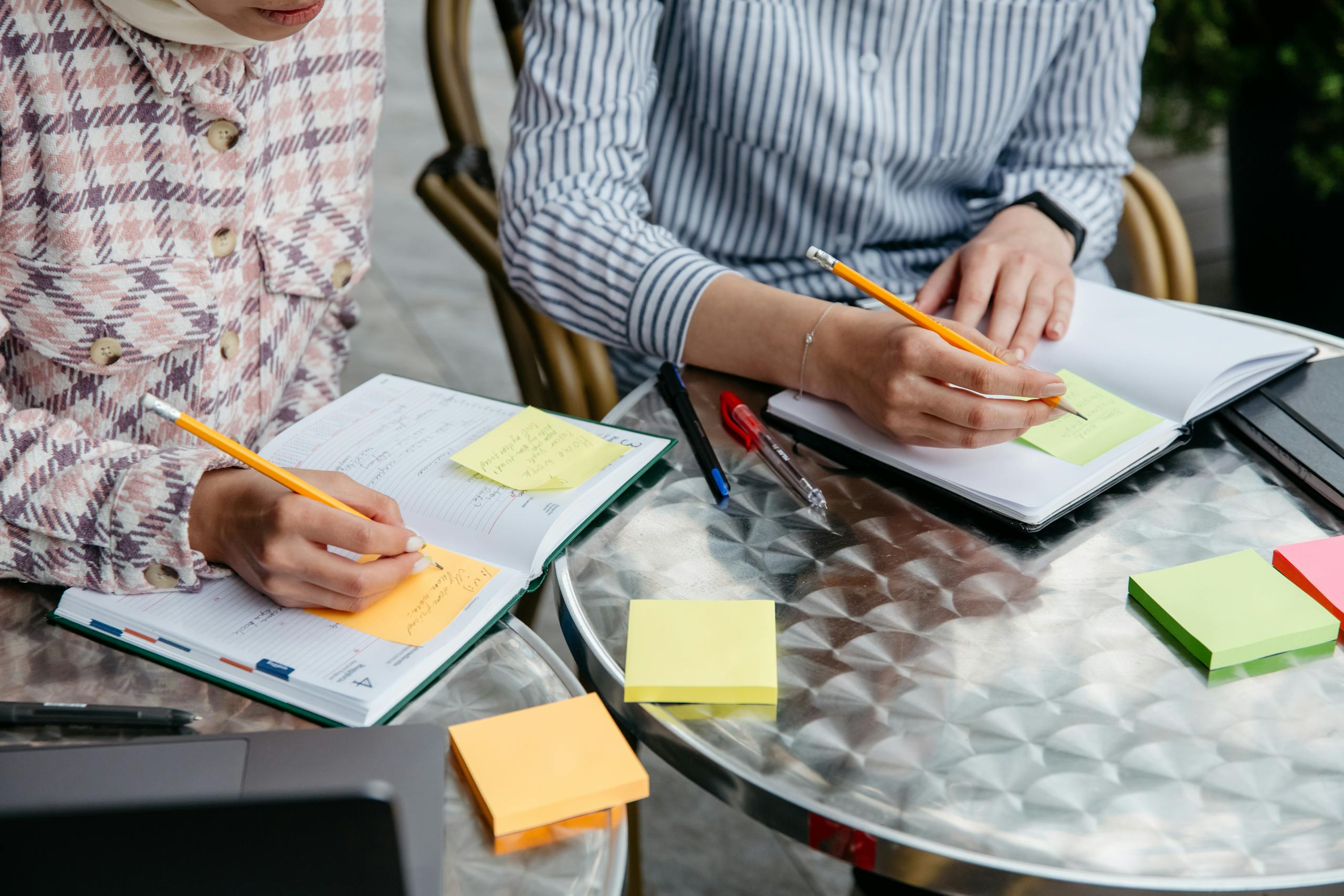 Two women writing notes in notebooks with sticky notes and pens on a table.