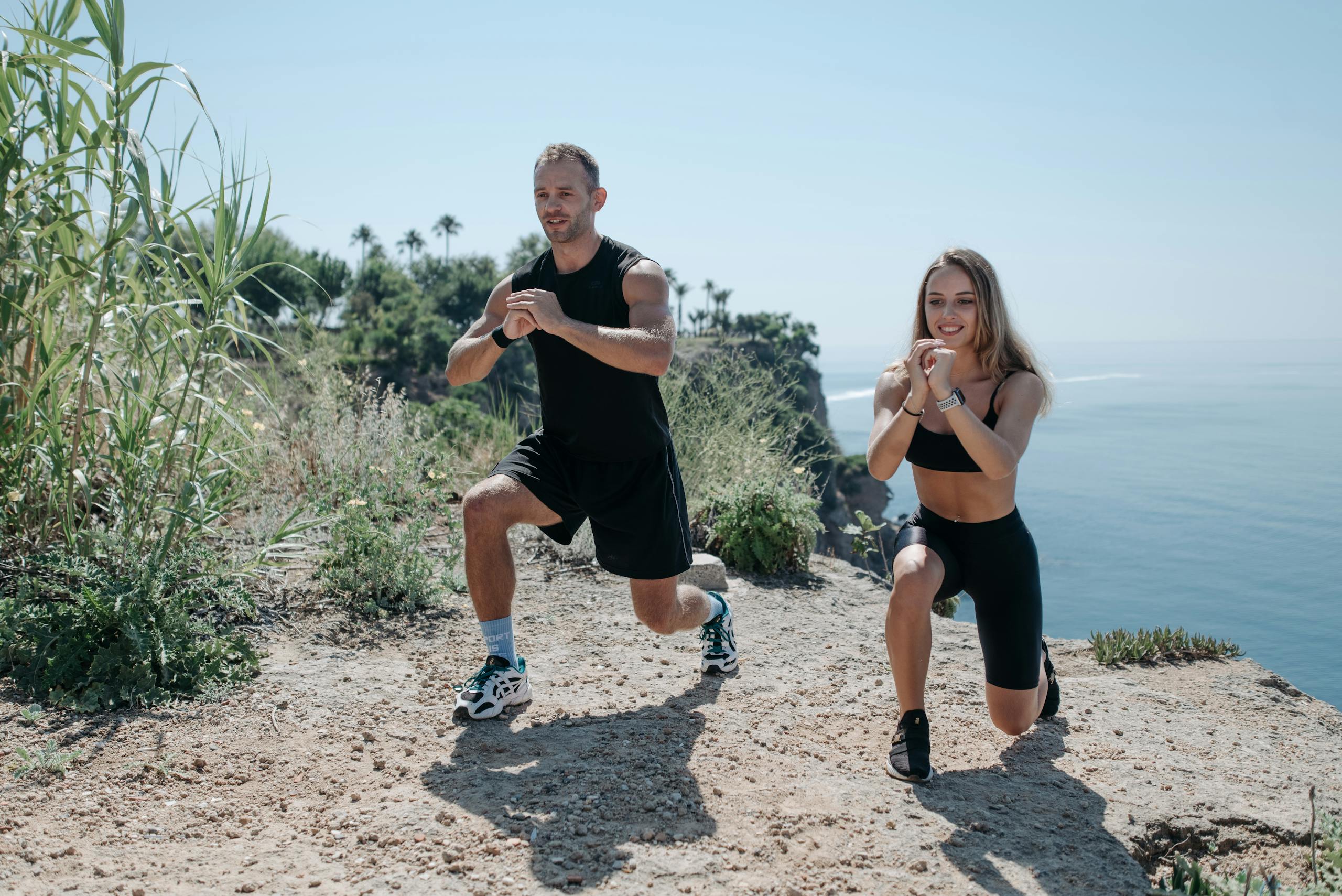 A man and woman exercising on a rocky cliff overlooking the ocean on a sunny day.