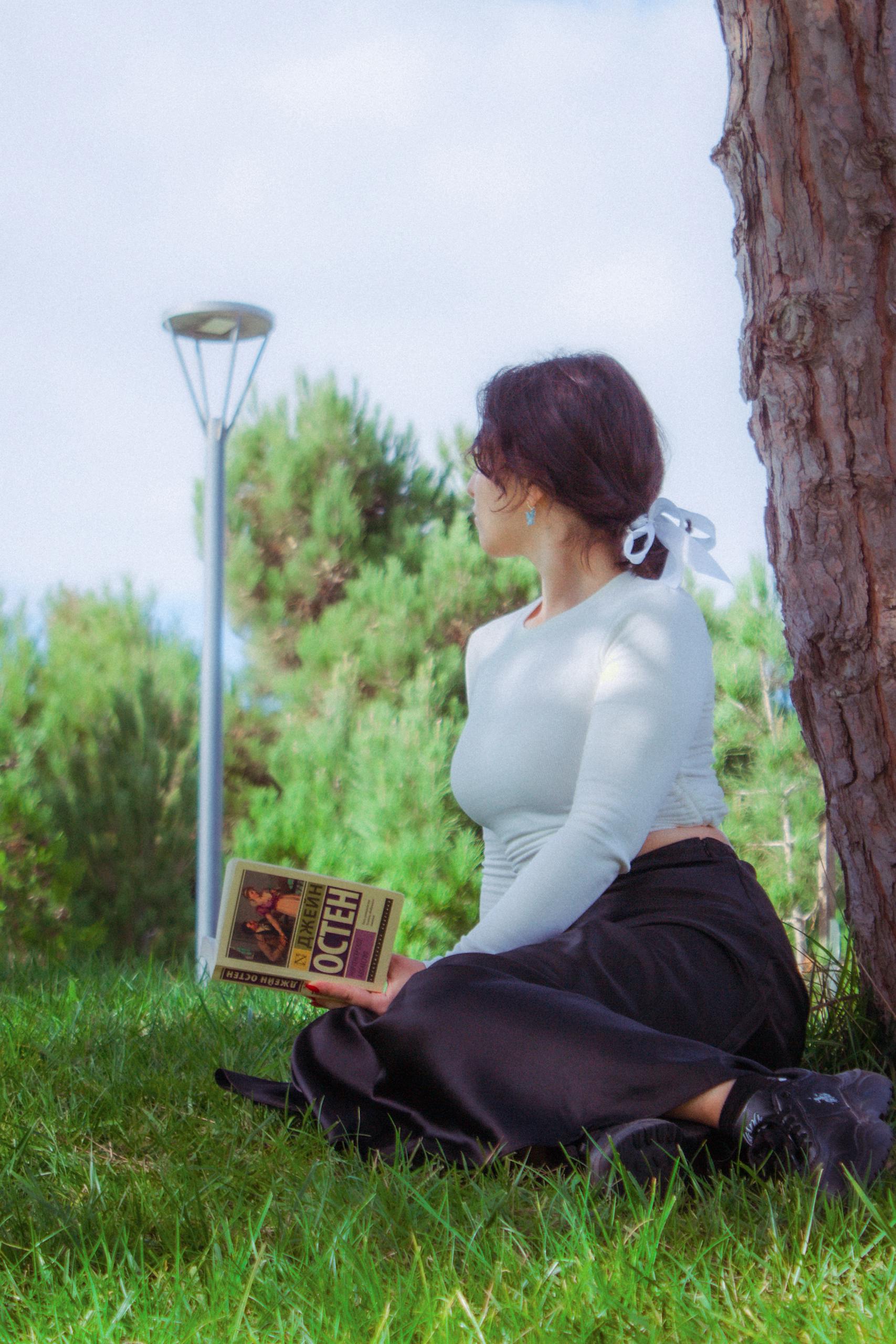 A woman sitting on grass in a park reading a book on a sunny day.