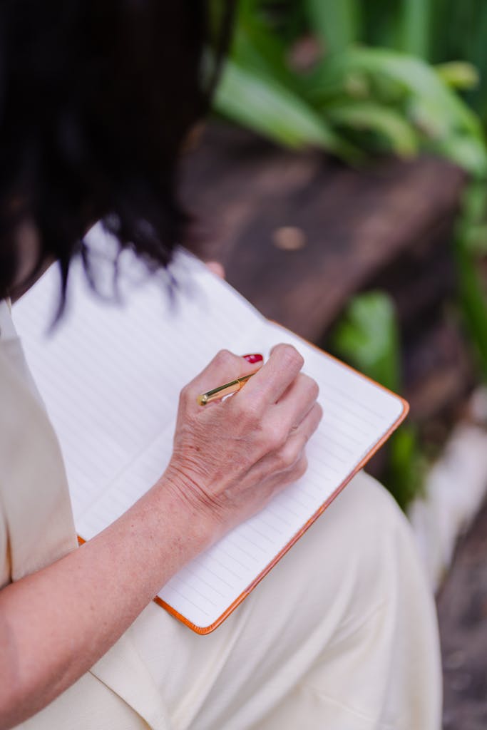 A woman writing in a lined journal while sitting outdoors in a garden.