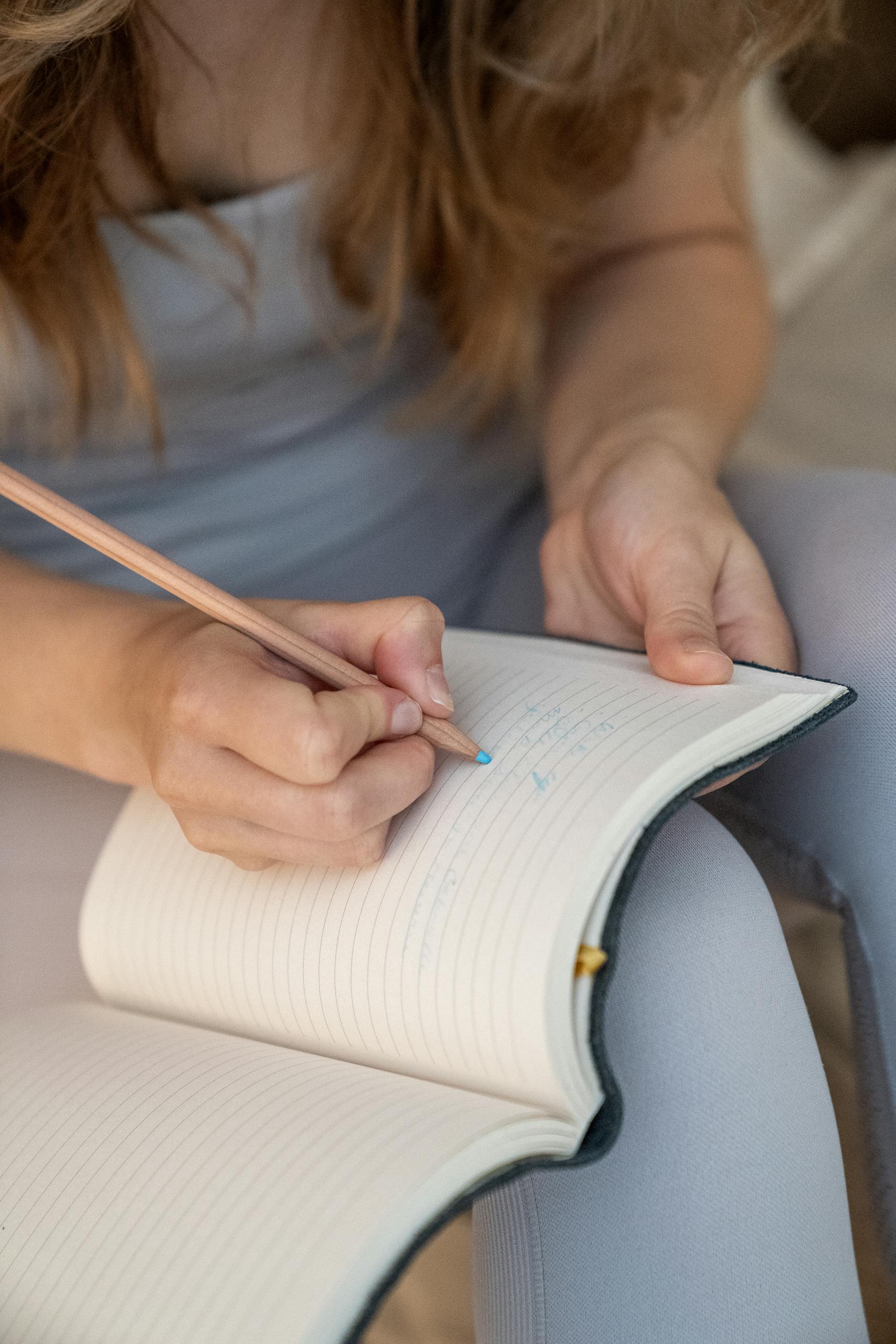 A woman writing in a lined notebook with a pencil, creating notes or journaling.