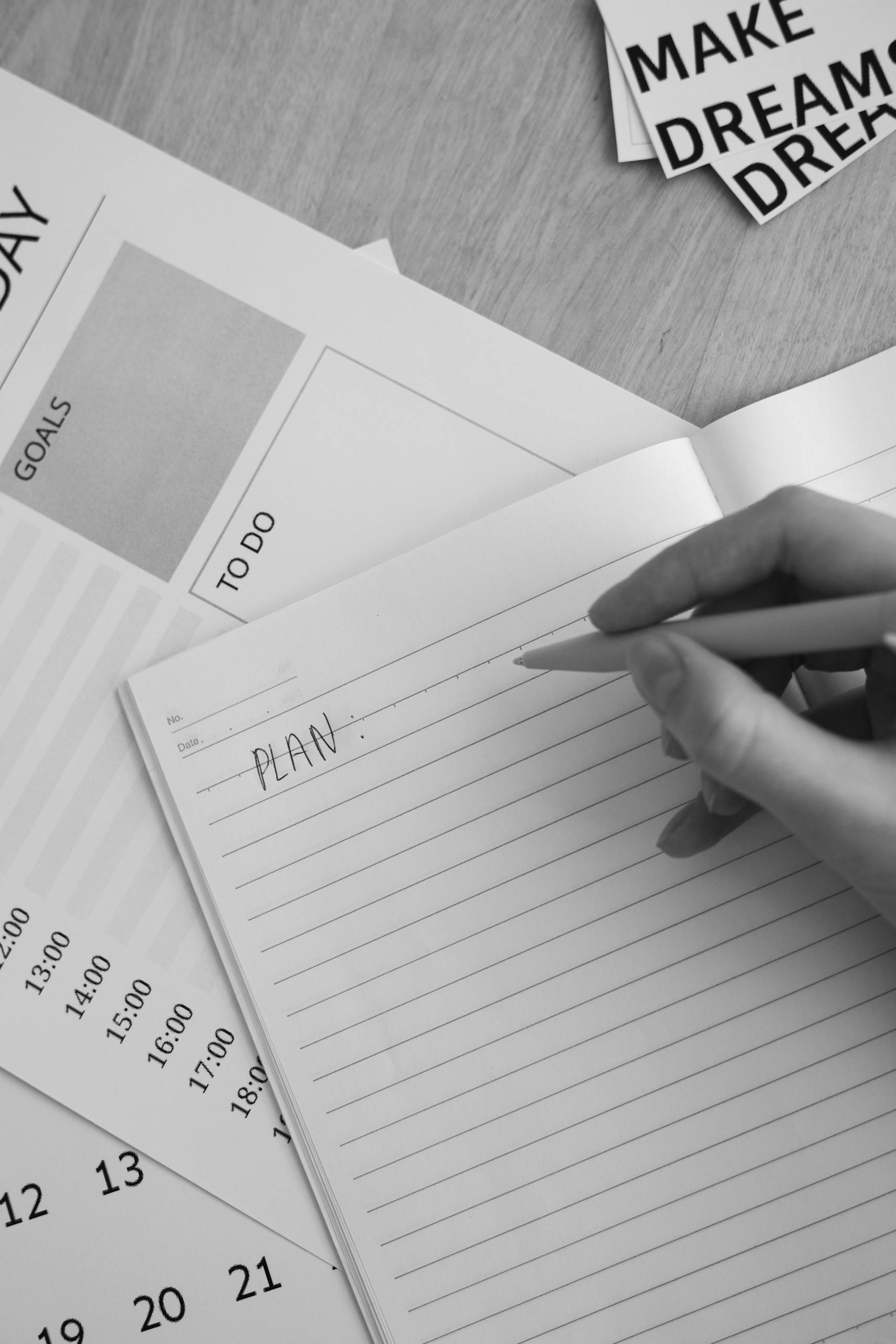 Black and white photo of hand writing 'plan' in notebook with planning materials around.