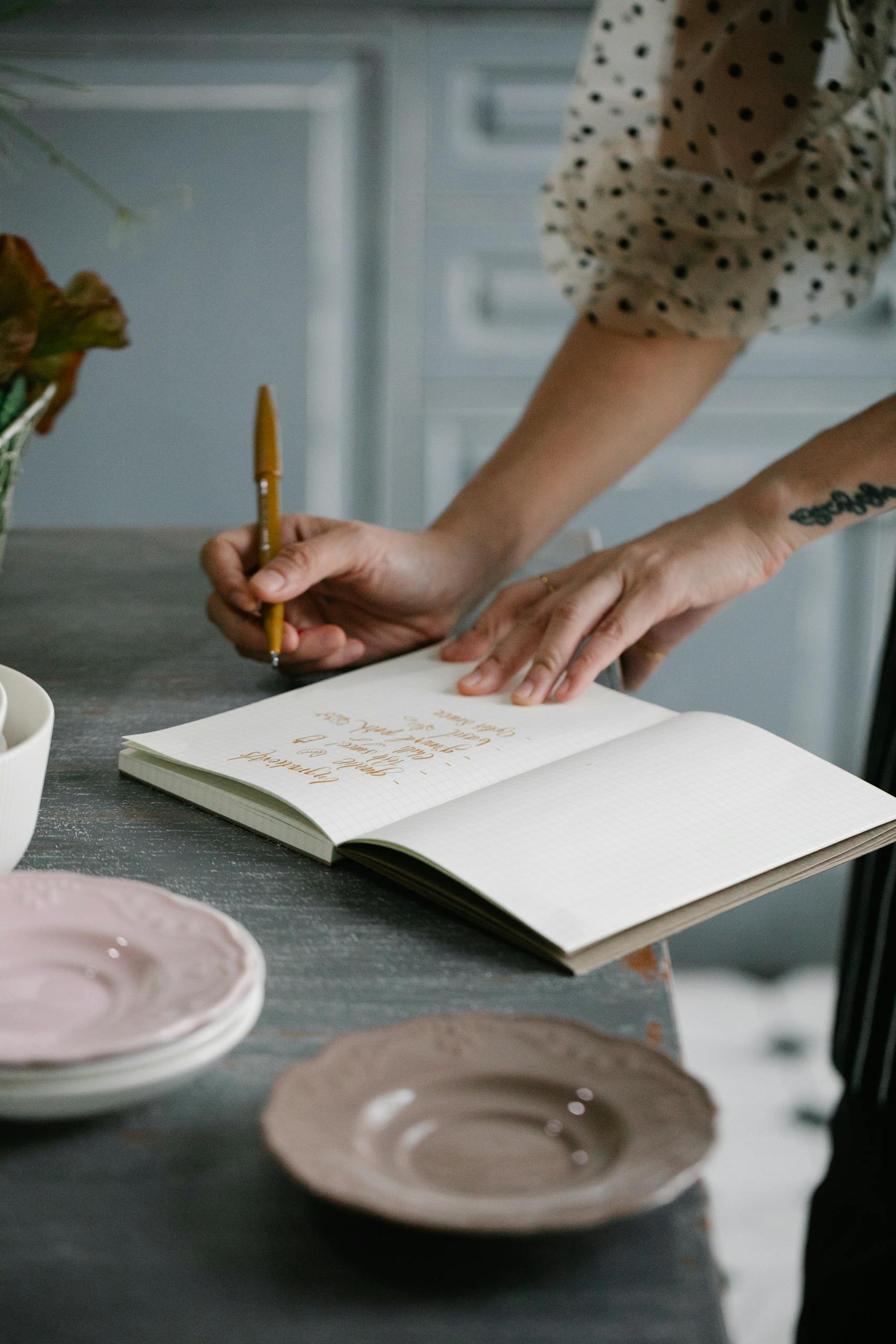 Close-up of a woman writing in a notebook at a table set with plates in a kitchen setting.