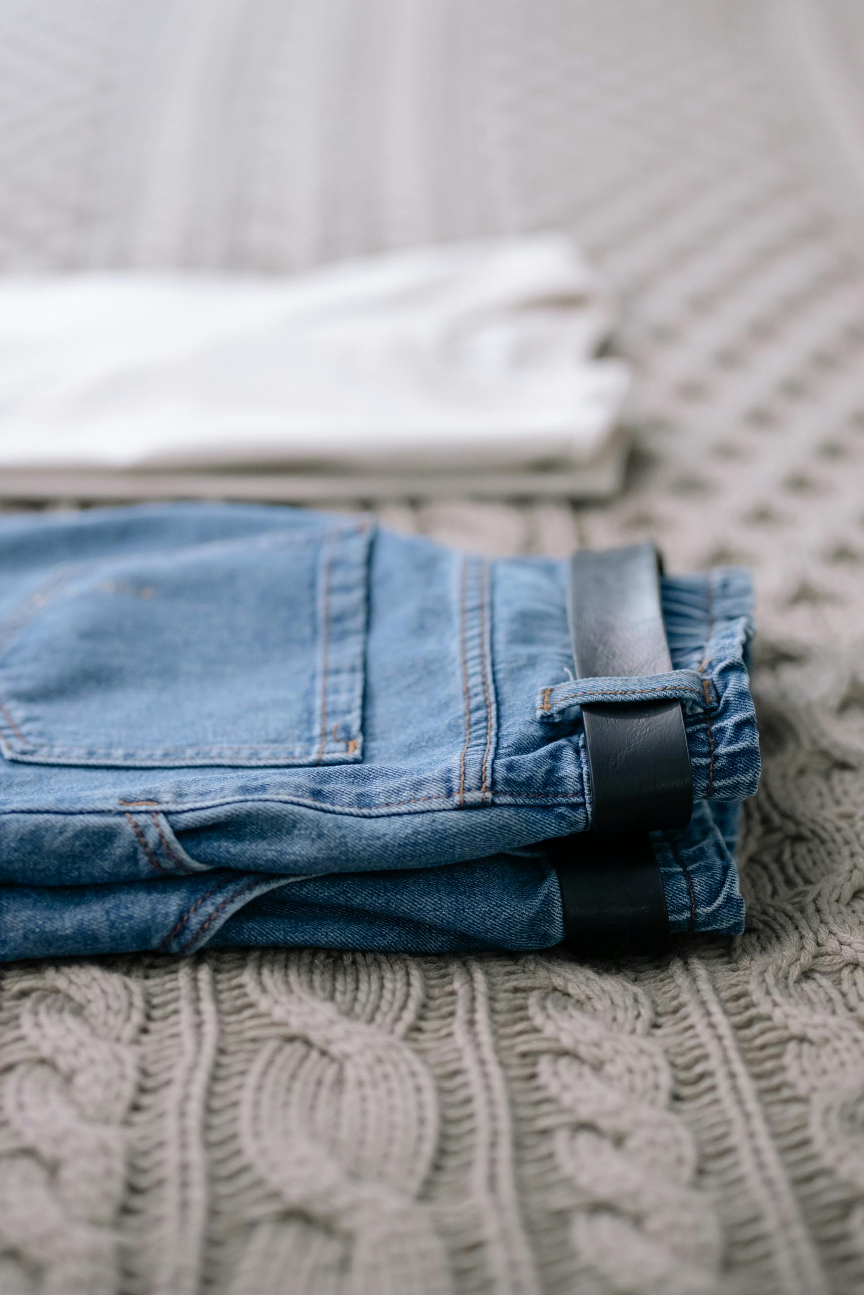 Close-up of folded jeans with belt on a textured knit blanket, showcasing fabric details.