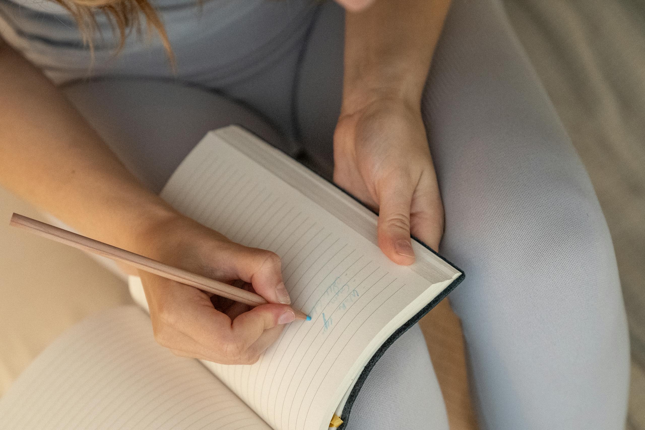 Close-up of hands writing in a lined notebook with a pencil. Soft and serene indoor setting.