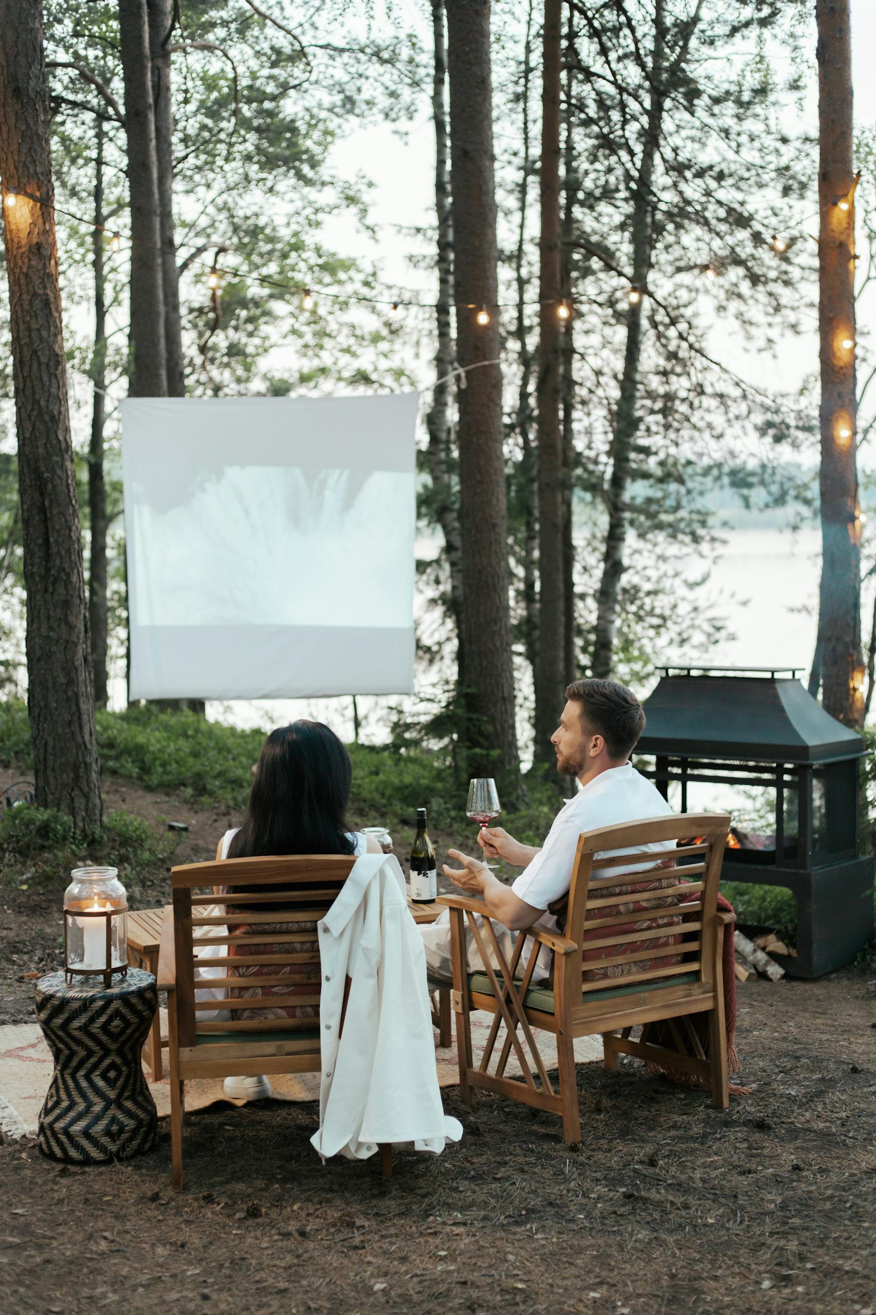 Couple enjoys a cozy outdoor movie night with wine under string lights.