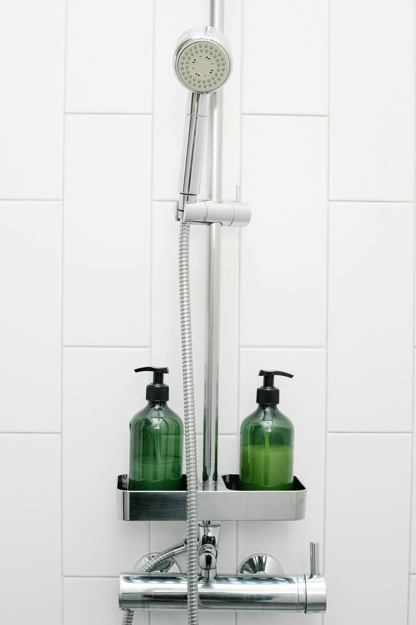 Handshower and hose attached to white tiled wall near bottles with essential skin care products in light bathroom