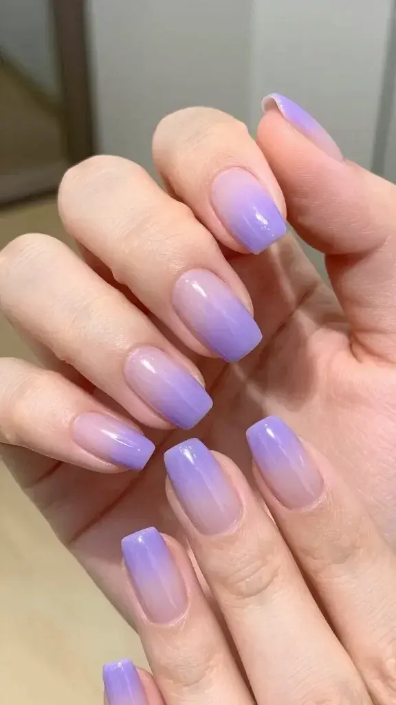 Create a hyper-realistic, high-resolution close-up portrait of a hand with lavender latte ombre nails as the main subject, styled to convey a soft, summery aesthetic. The nails should feature a smooth gradient transitioning from a deeper lavender at the tips to a pale lilac near the cuticles, with a sheer lilac base. Add a delicate, pearlescent sheen to the surface to mimic latte steam for depth and a subtle shimmer top coat for a gentle, wearable glow. The lighting should be soft and natural, with warm ambient tones suggesting a sunny, office-friendly vibe. The background should be softly blurred in pastel tones to keep attention on the nails, and the hand should be posed gracefully to highlight nail shape and gradient. The overall image should feel polished, chic, and approachable. No text overlays.