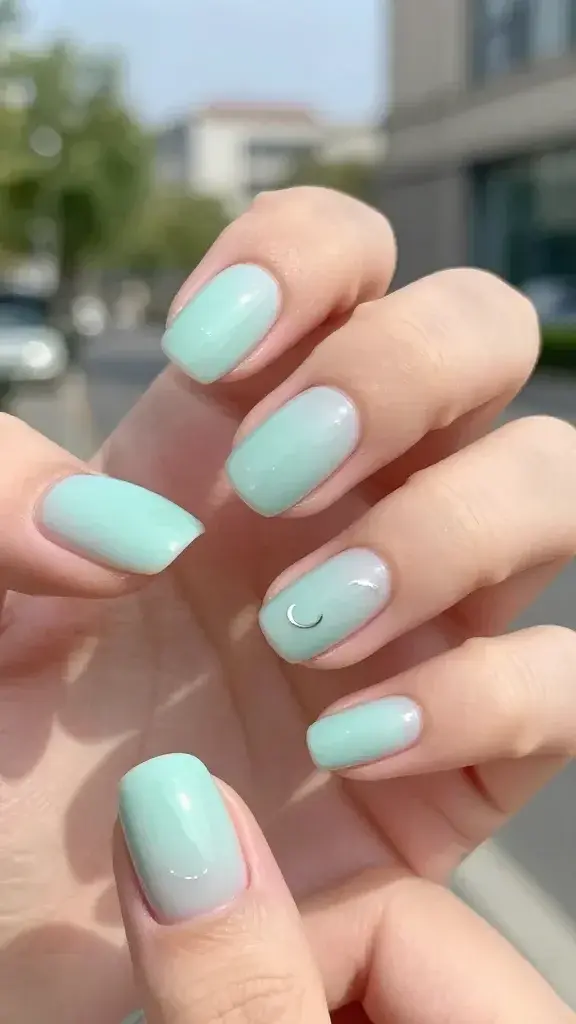 A hyper-realistic close-up photograph of a hand with pale mint nails, each nail featuring tiny silver crescent moons catching the light. The background is softly blurred outdoor daylight with a cool, calming vibe. The nails have a ultra-glossy glassy finish, reflecting subtle highlights and tiny sparkles from overhead sun. The overall look is minimal yet offbeat, with a clean manicure on well-groomed nails, and a hint of natural greenery in the out-of-focus background to emphasize a serene, celestial mint mood. No text or overlays.