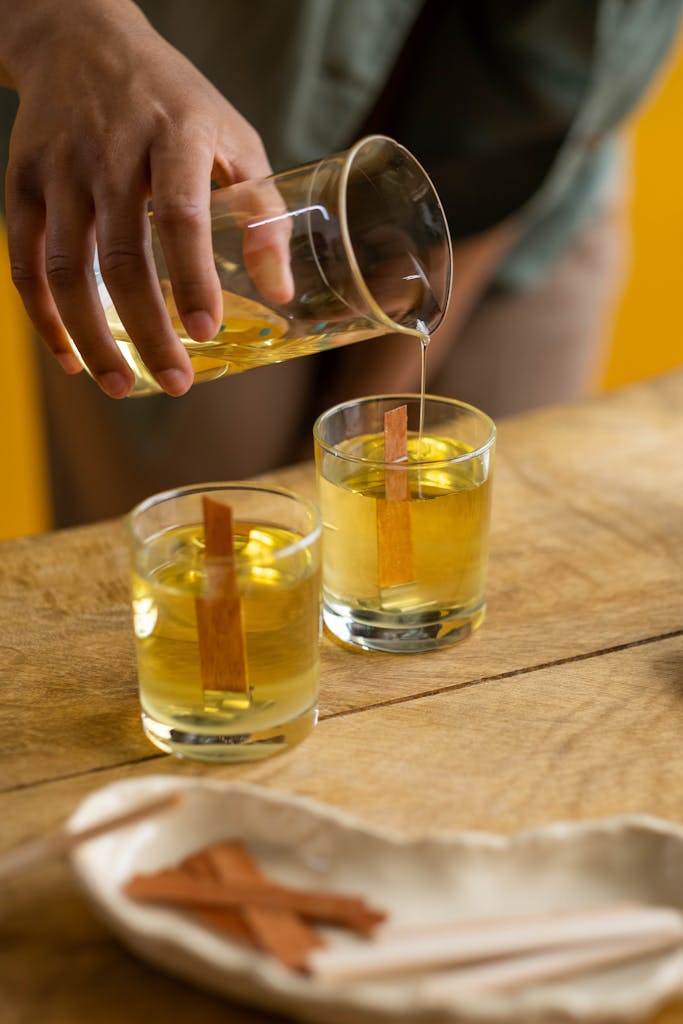 Person pouring yellow wax into glasses with wooden wicks on a wooden table.