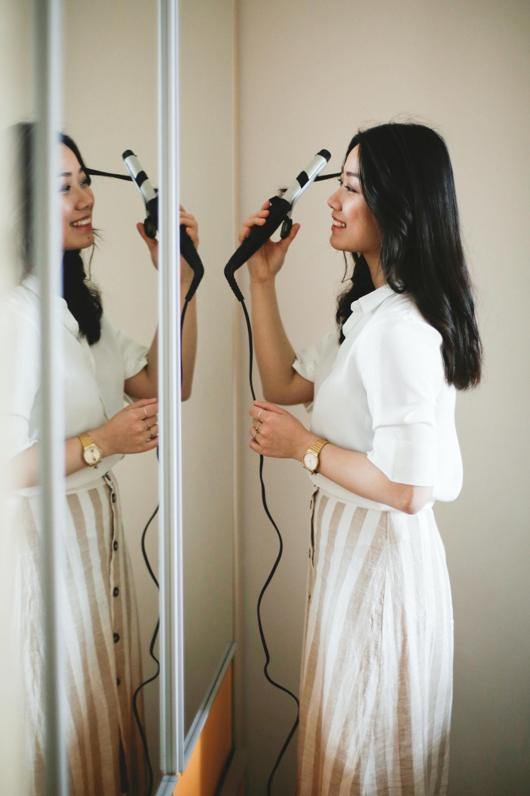 Side view of happy Asian female smiling and looking at mirror while coiling hair in morning at home