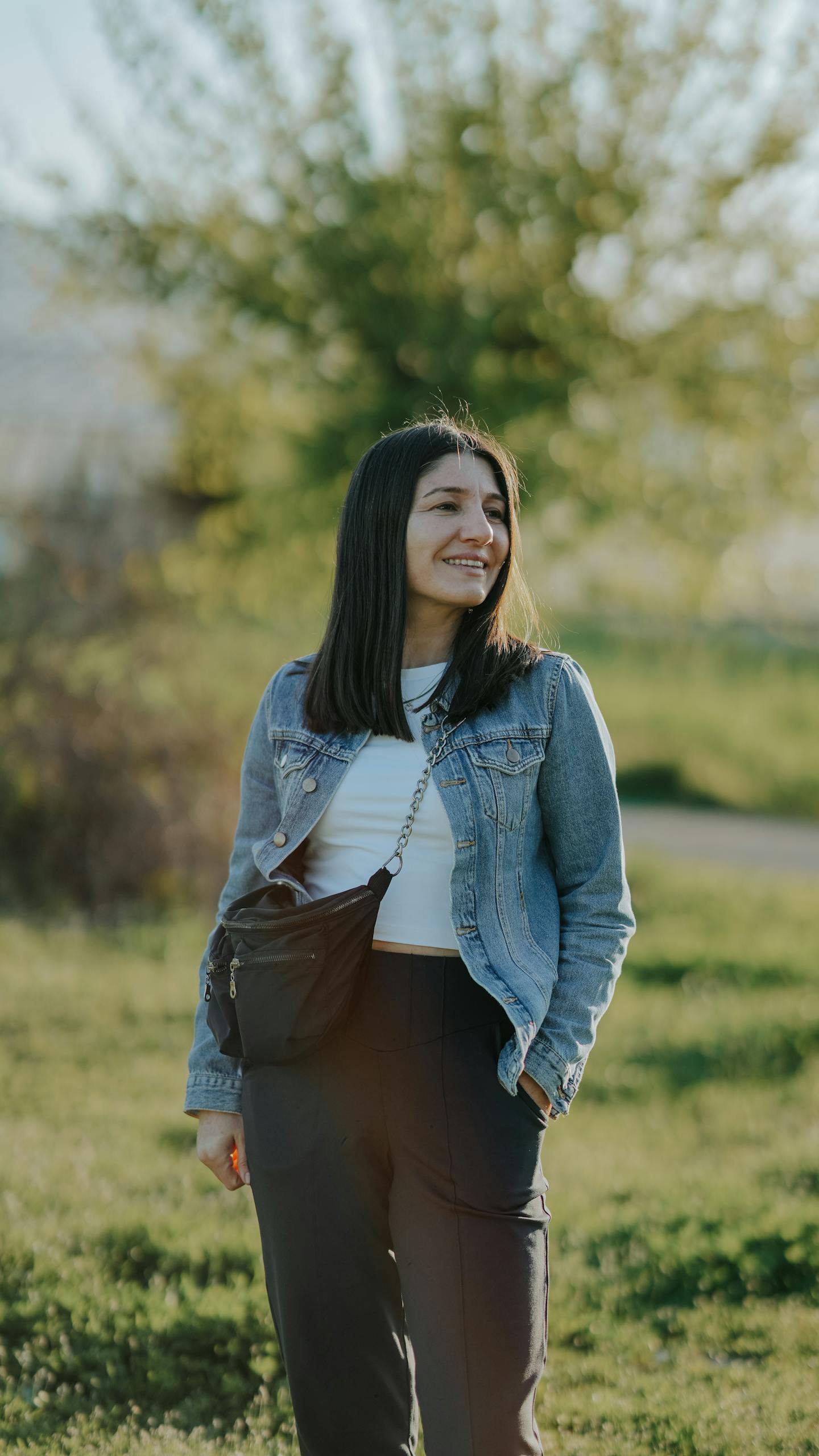 Smiling woman in denim jacket enjoying a sunny day outdoors, surrounded by greenery.