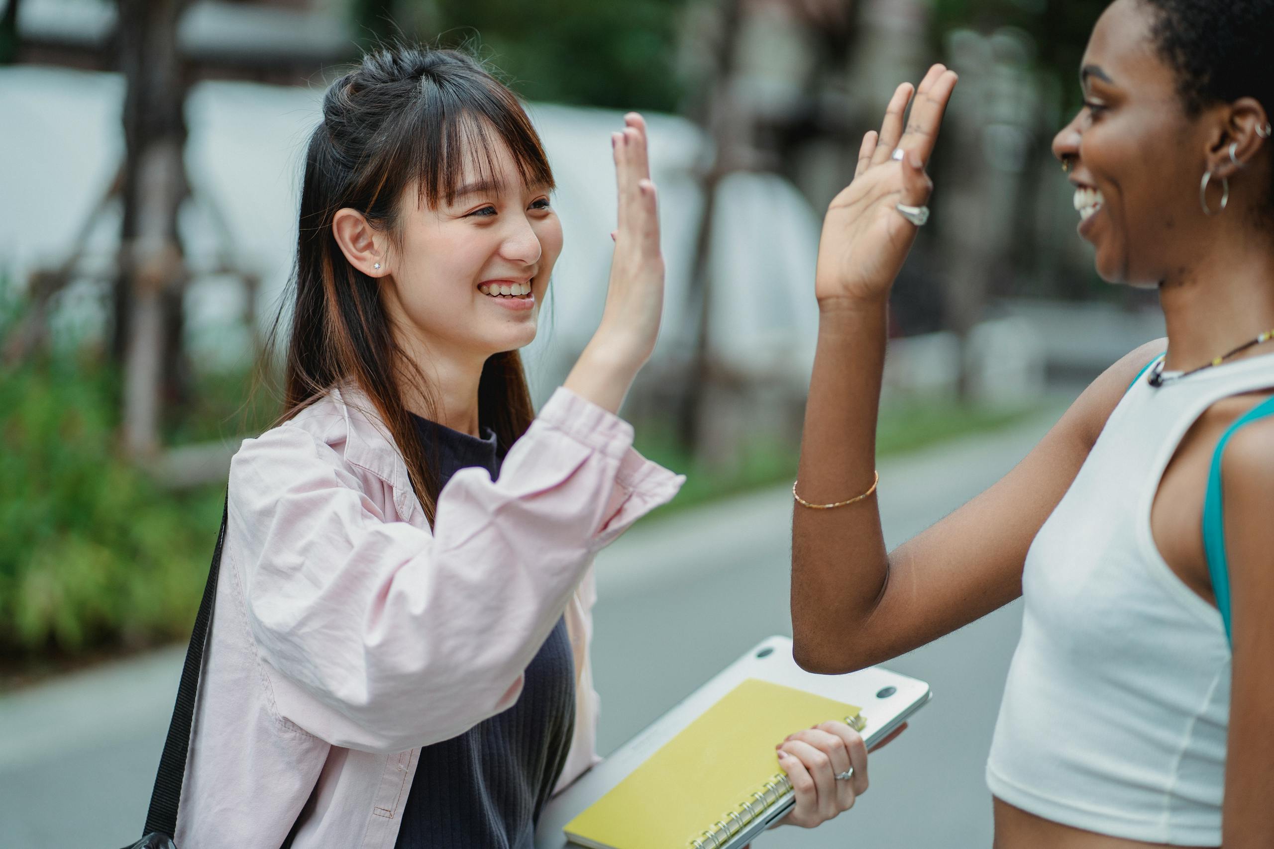 Smiling young multiracial women standing in middle of road with computer and notebook and giving high five while looking at each other