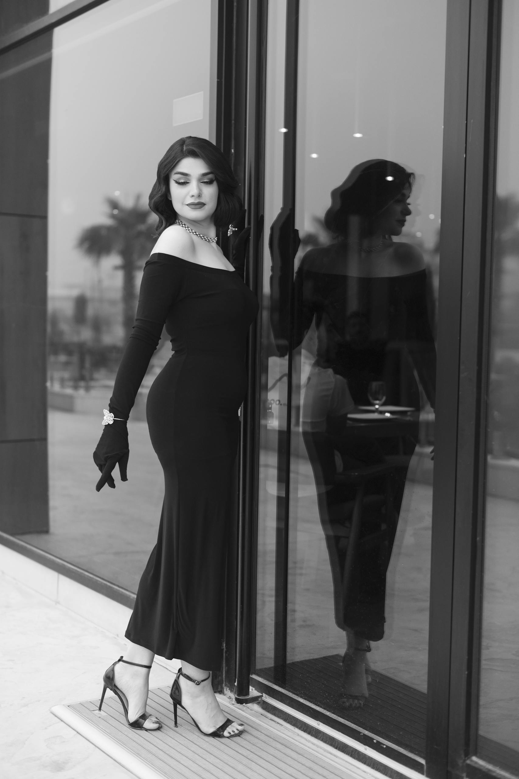 Black and white photo of an elegant woman in high fashion, standing near glass doors.