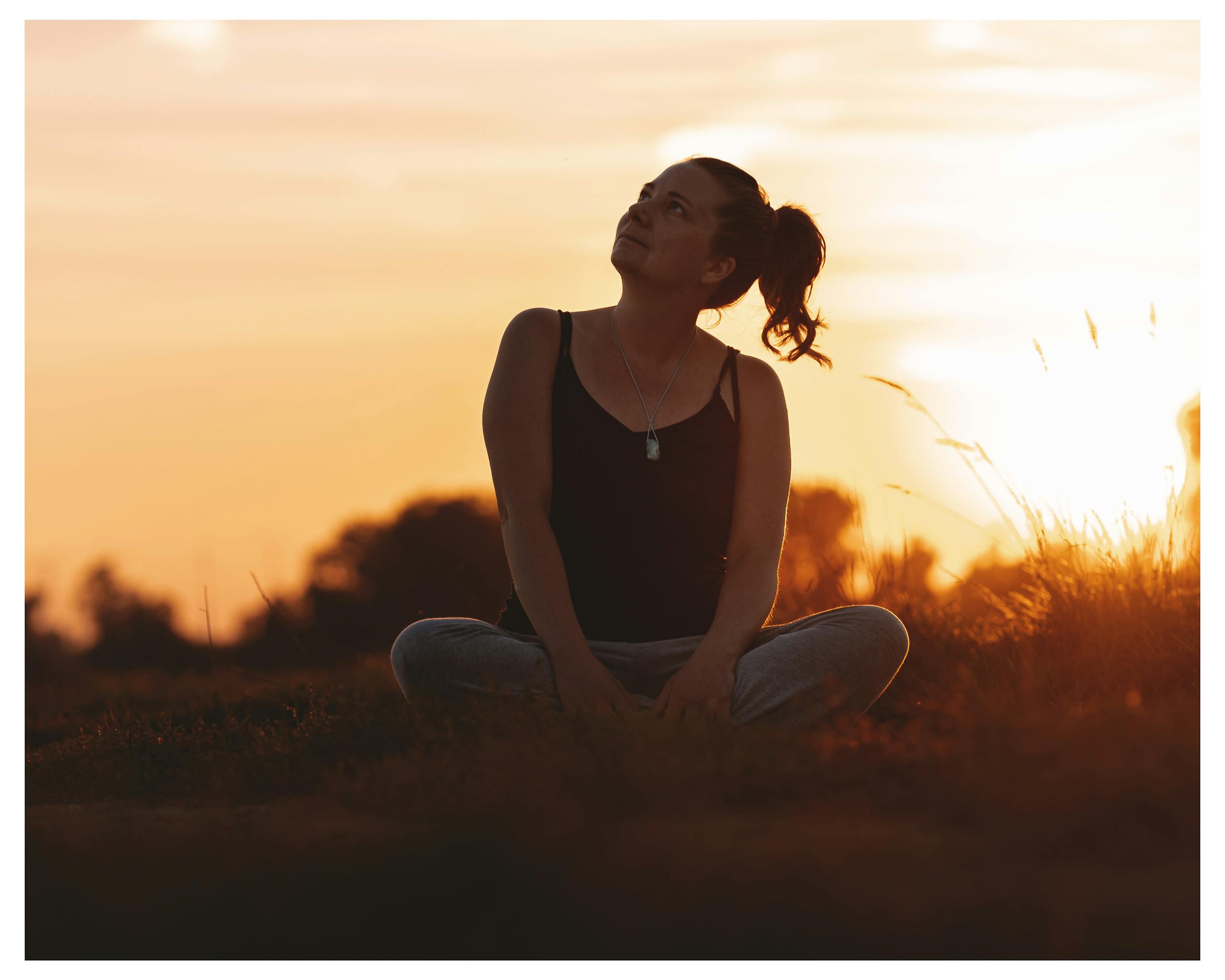Serene woman sitting in field, looking at sunset, wearing black tank top.