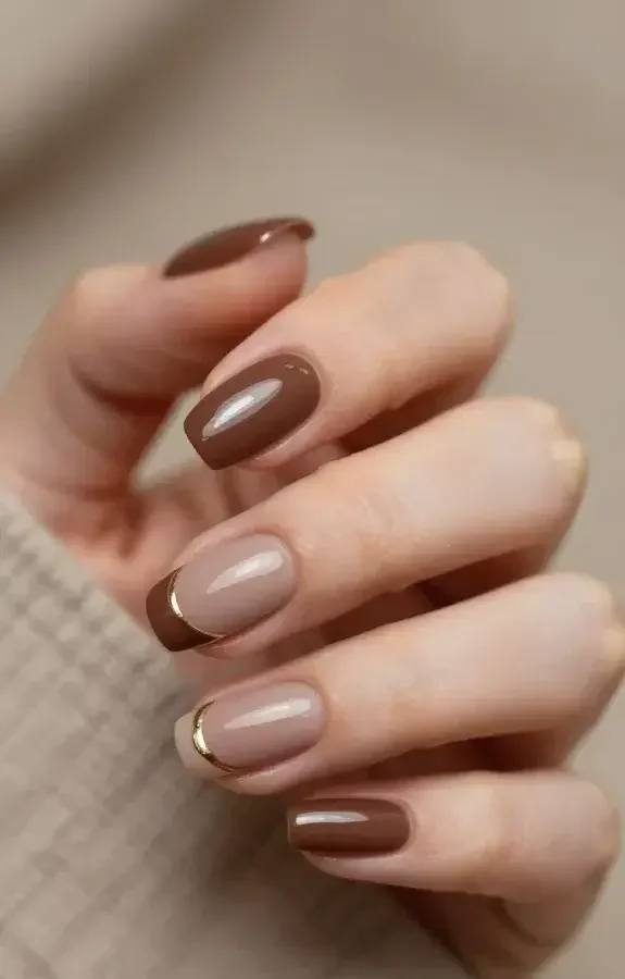 Create a hyper-realistic close-up photograph of a hand displaying a fall manicure titled "Cozy Cocoa Neutrals With a Twist." The main subject is a well-manicured hand resting on a soft, neutral-toned fabric backdrop with warm ambient lighting. Focus on one hand held slightly at an angle to reveal the nails clearly. Nail design features: base colors in deep cocoa or taupe nude with a subtle, modern twist; a single thin champagne stripe or dot placed near the cuticle on each nail as a delicate accent. The finish is a glossy, museum-polish shine that catches the light and reflects softly. The nails are medium-length with an almond or oval shape, evenly polished, with clean edges. The cuticles are neat, and the skin around the nails appears hydrated. Include a shallow depth of field so the nails are in crisp focus while the background blurs gently. No text or overlays in the image.