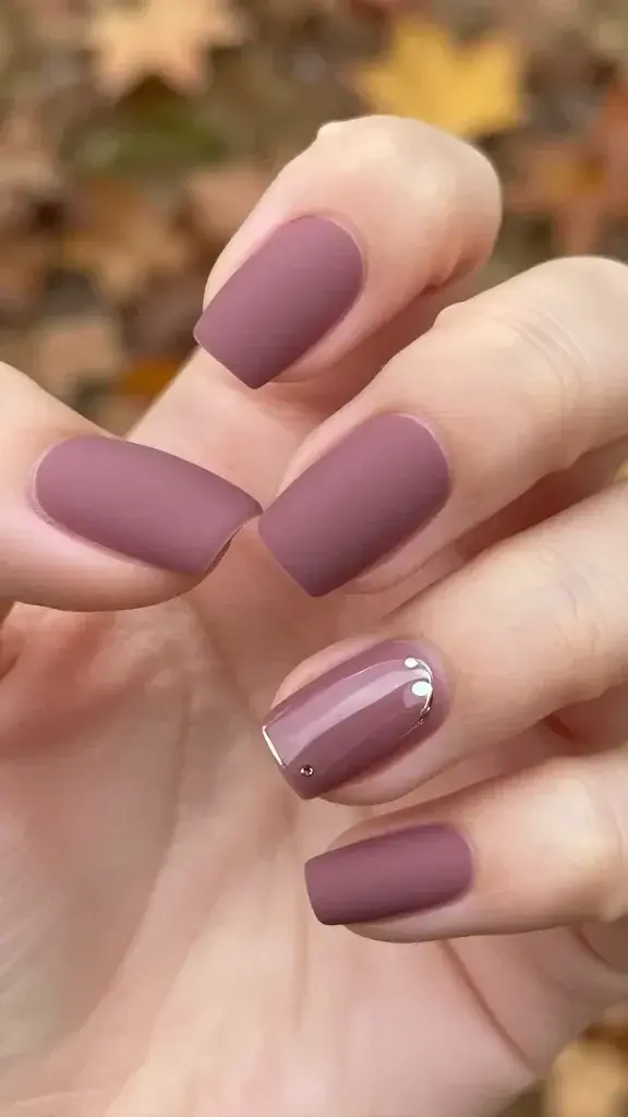 A high-resolution, close-up portrait-style shot of a hand with fall-inspired velvet matte mauve nails, showcased top-to-fingertips against a soft, blurred autumn background. The dominant nails wear a velvety, dusty mauve matte finish with a single glossy accent: one nail on each hand has a tiny metallic dot or a delicate metallic line, positioned subtly near the cuticle or along one edge. The rest of the nails maintain a smooth, rich matte texture. Lighting is warm and soft, highlighting the contrast between the velvety matte nails and the small glossy accent to make the nails pop in photos. The hand is posed naturally, slightly curled to emphasize nail shape (squoval) and symmetry, with a minimal, non-distracting manicure environment. No text or overlays.