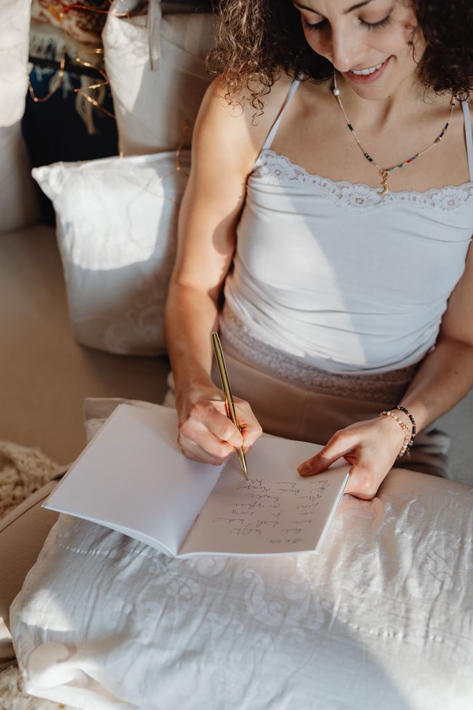A brunette woman writes and smiles while sitting comfortably indoors with a notebook.