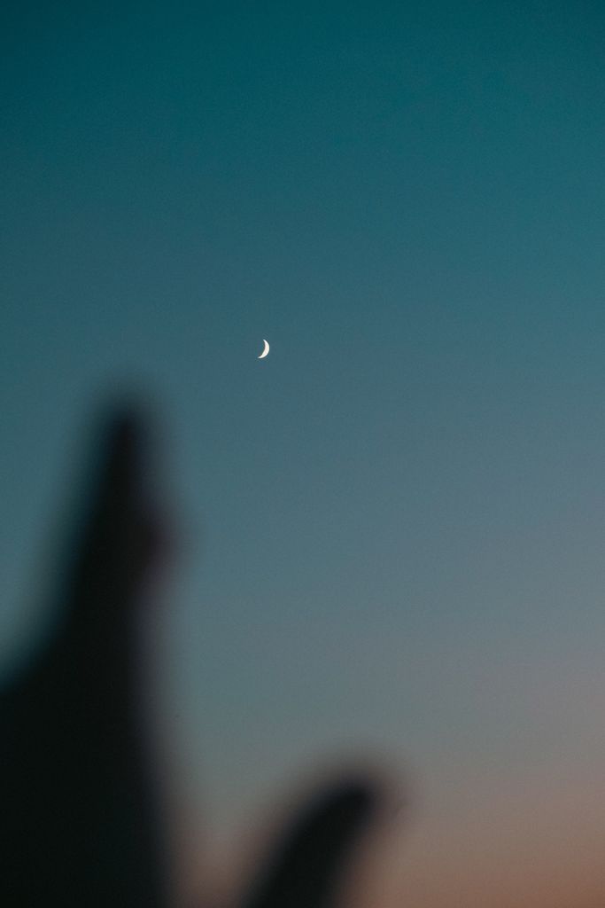 A crescent moon in a twilight sky with silhouetted hands creates a serene ambient scene.new moon manifestation