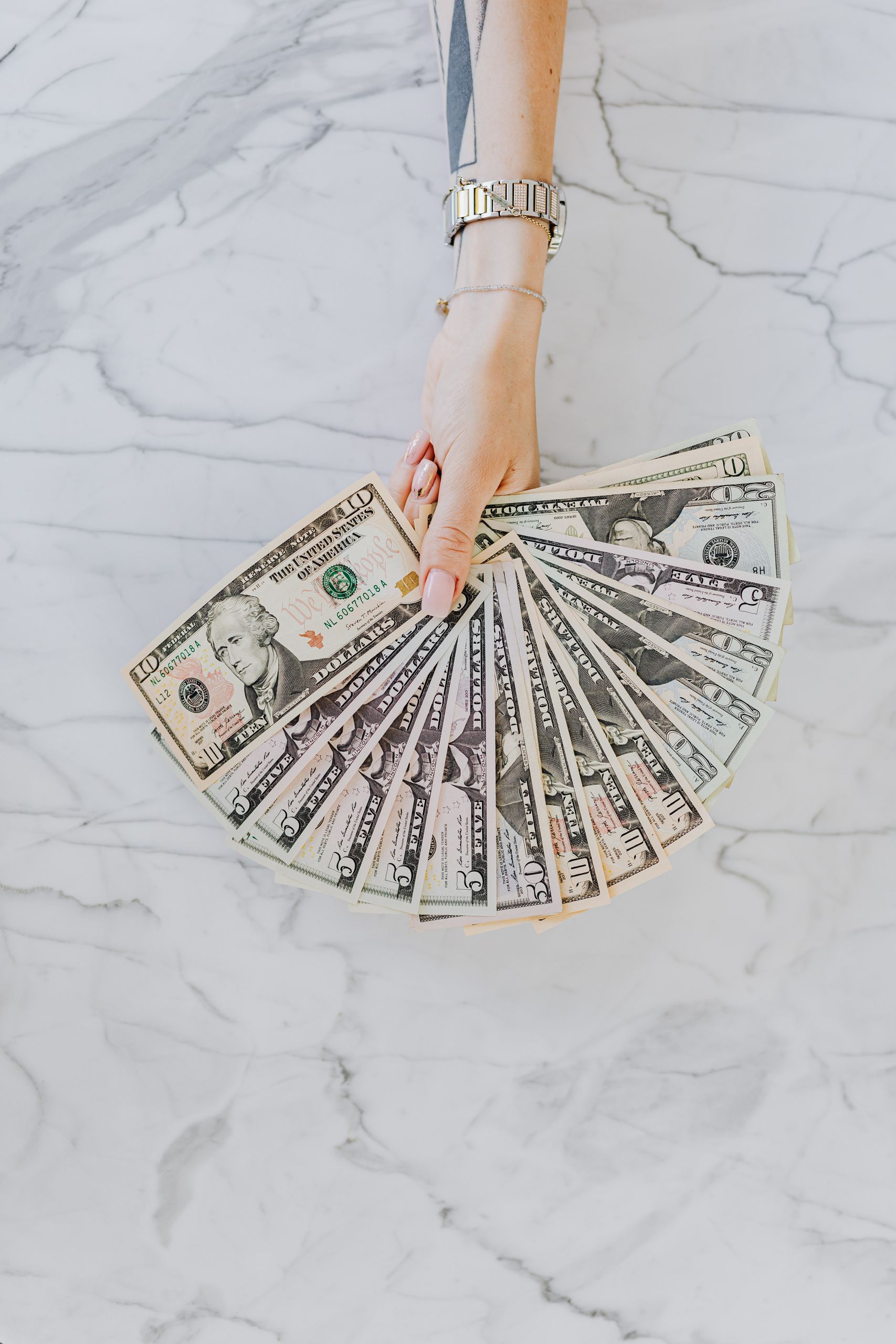 A hand with wristwatch holding a fan of US dollar bills on a marble surface, symbolizing wealth and financial abundance.