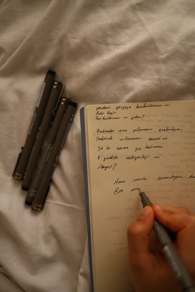 A hand writes in a notebook with pens nearby on a cozy bed setting.year-end reflection questions