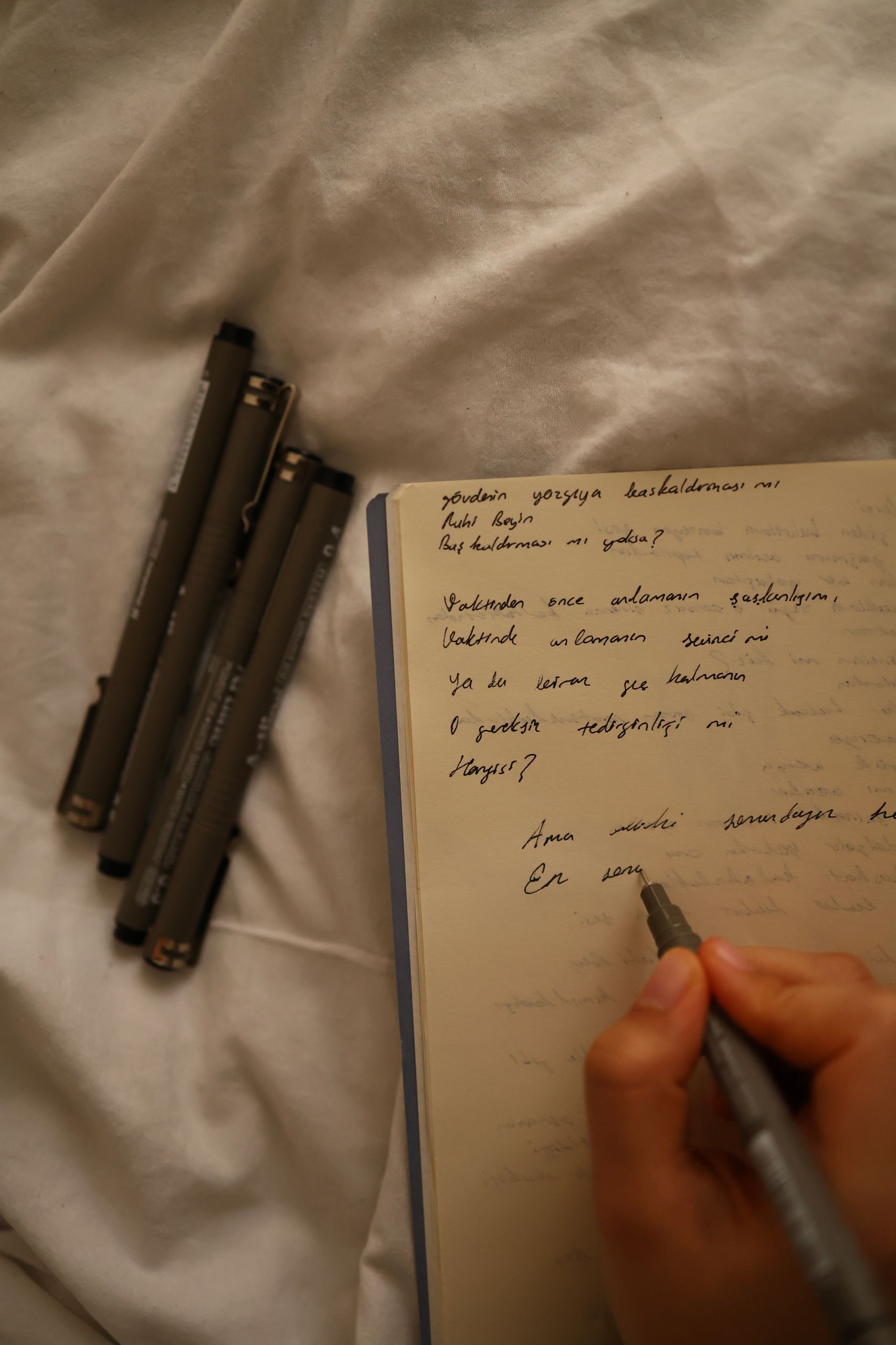 A hand writes in a notebook with pens nearby on a cozy bed setting.