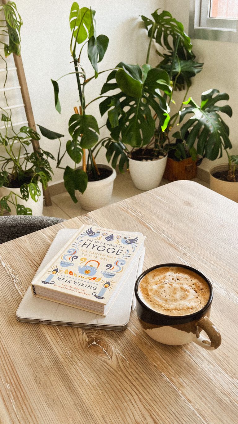 A hygge-inspired scene with a book, warm coffee, and lush indoor plants on a wooden table.