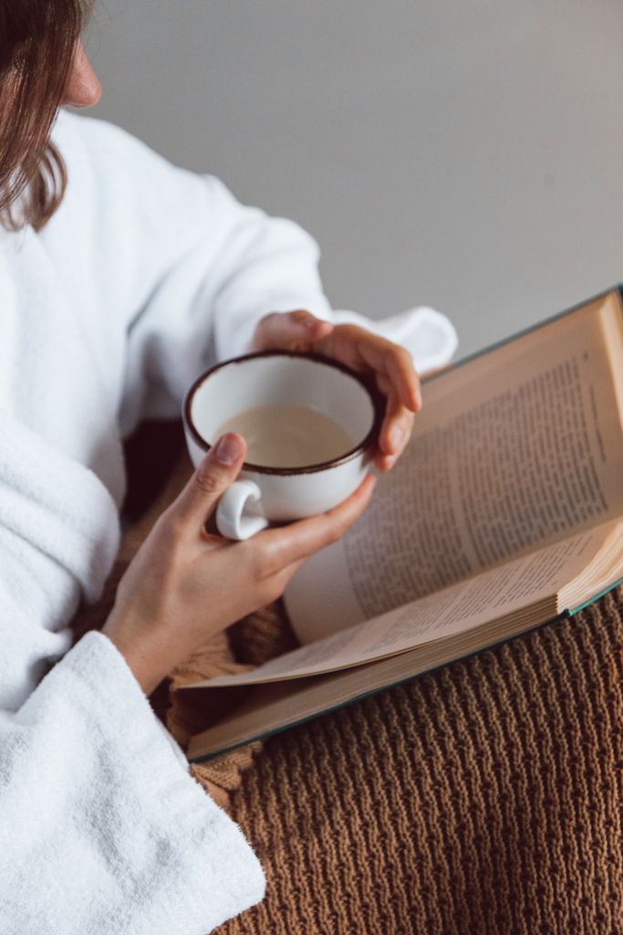 A person enjoying a cozy moment with a book and a cup of tea in hand, wearing a robe.best glow up tips