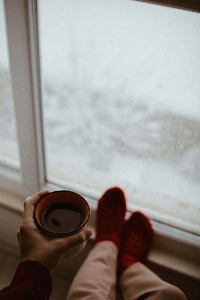 A person relaxes by a window, sipping coffee, wearing red socks, while enjoying a snowy view winter hygge.