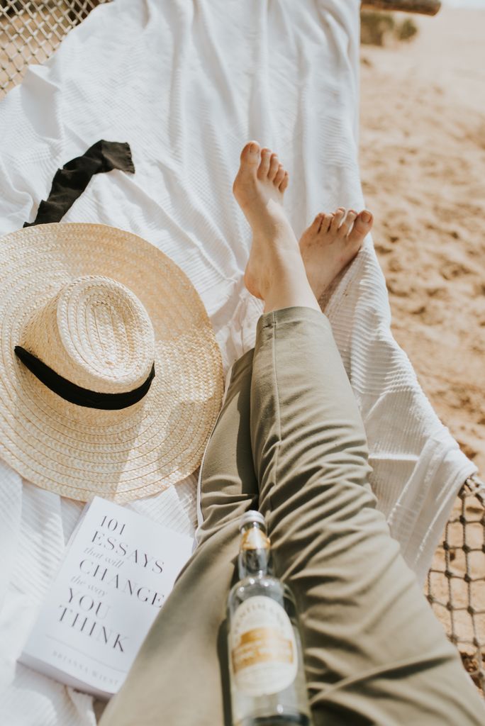 A person relaxing on a sunny beach with a straw hat, book, and drink, enjoying leisure time.