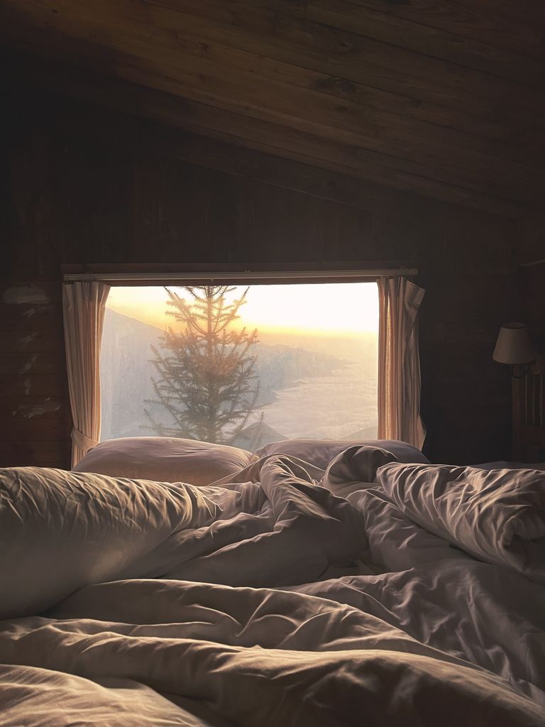 A serene bedroom with a view of sunrise through a large window in a rustic cabin.