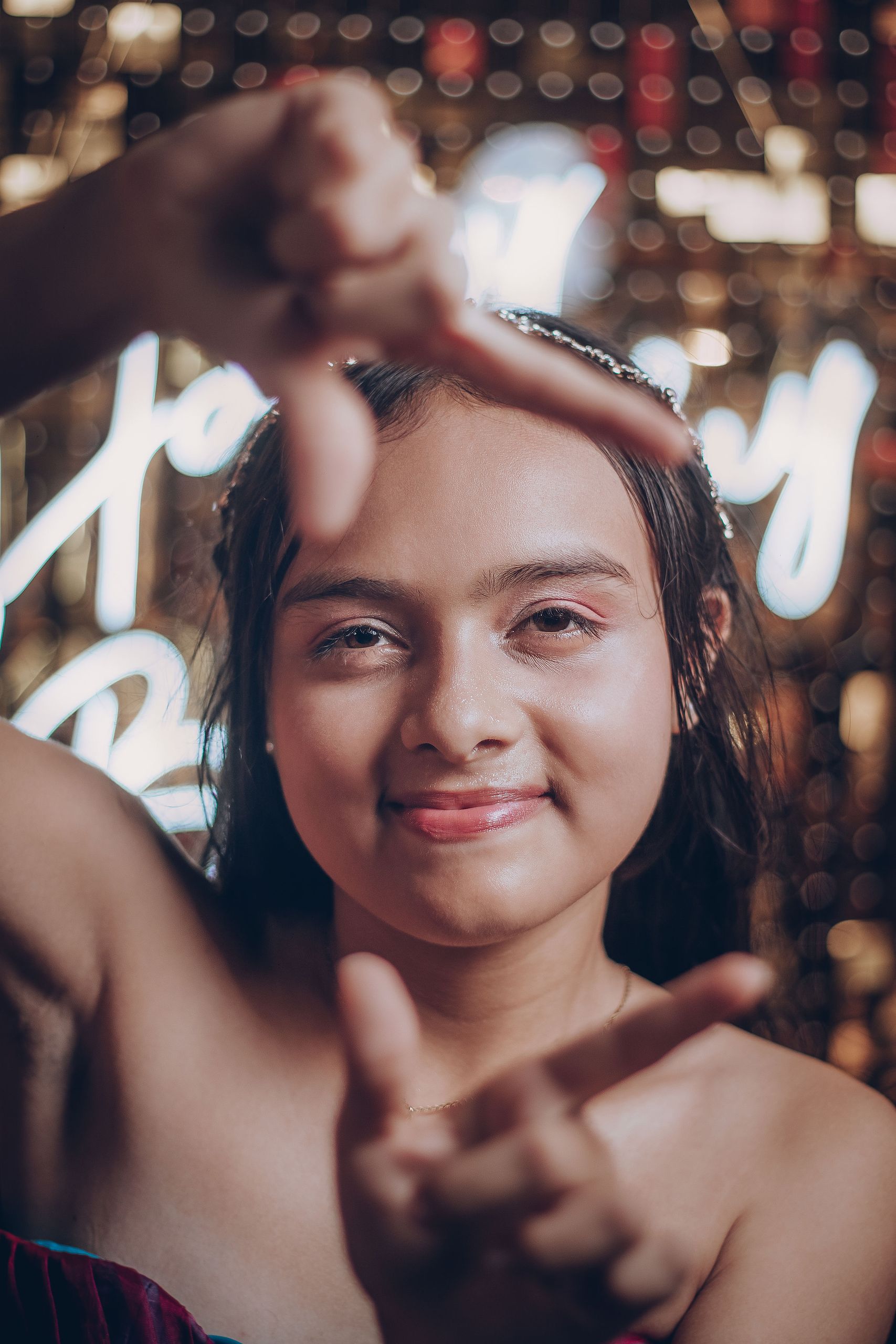 A smiling girl celebrating her birthday against a bokeh backdrop, framing her face with her hands.