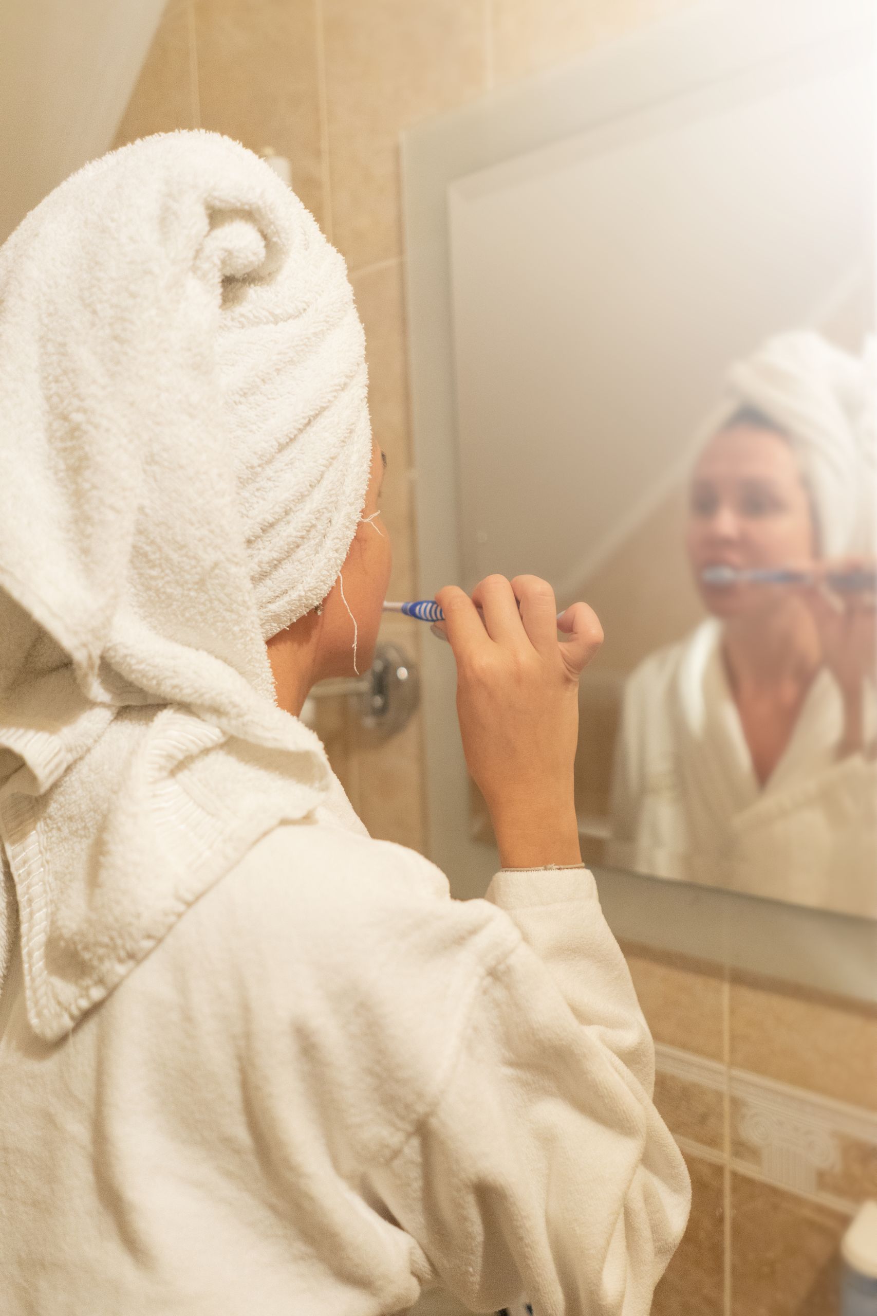 A woman in a bathrobe brushes her teeth while looking in the bathroom mirror, promoting daily hygiene.