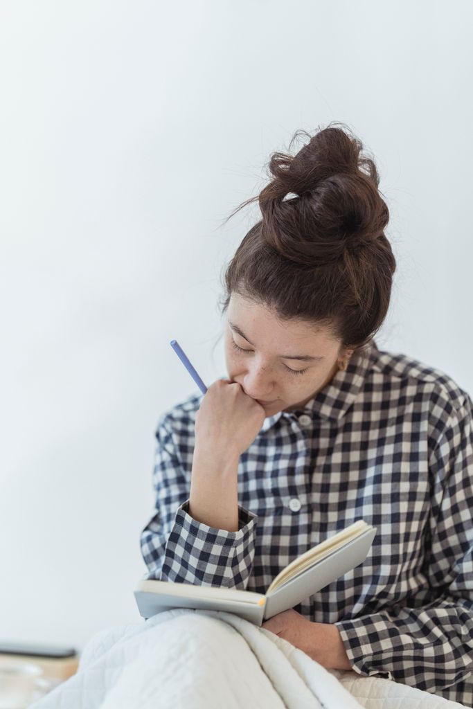 A woman in checkered shirt writes thoughtfully in a notebook indoors.