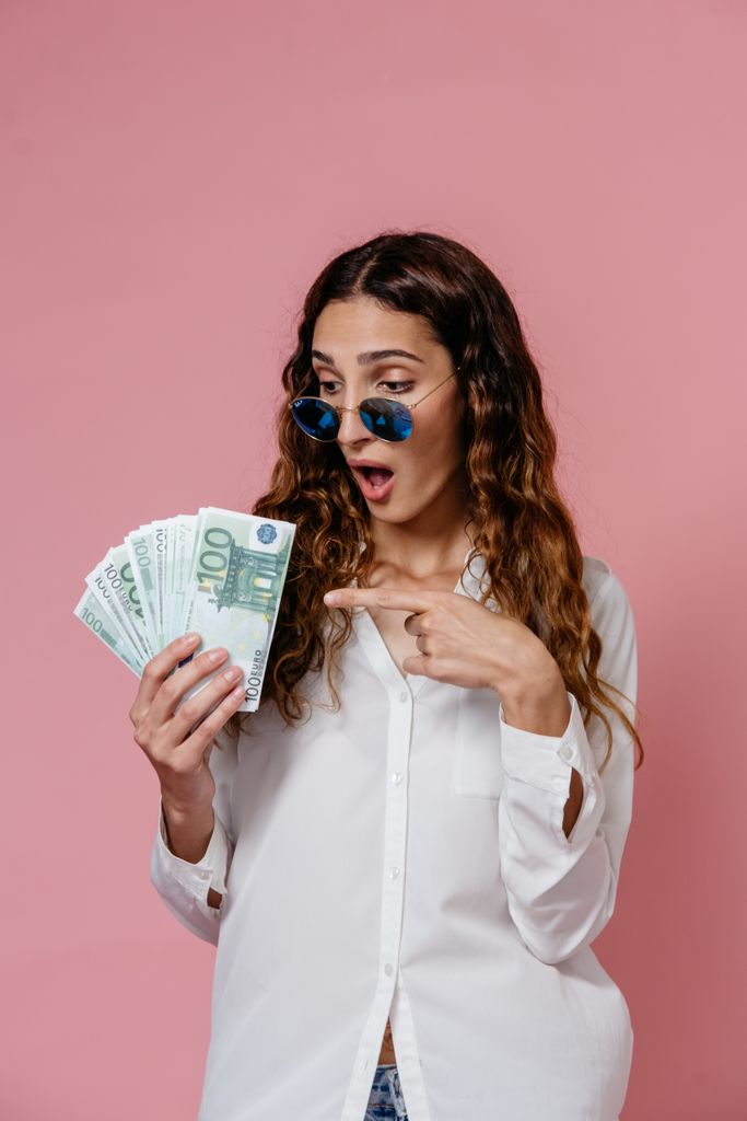 A woman in sunglasses holds and points at euro banknotes, on a pink background.