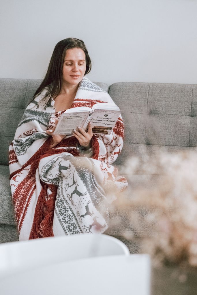 A woman sits comfortably on a sofa reading a book wrapped in a festive blanket.