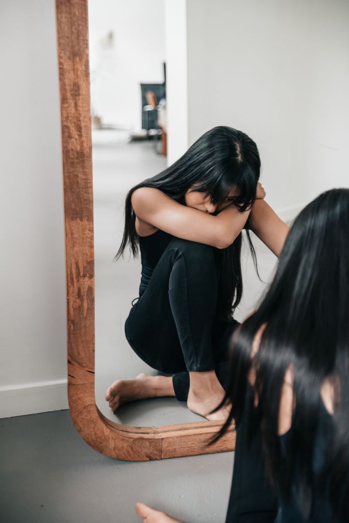 A woman sitting pensively by a large wooden mirror indoors, reflecting on her thoughts.how to reinvent yourself