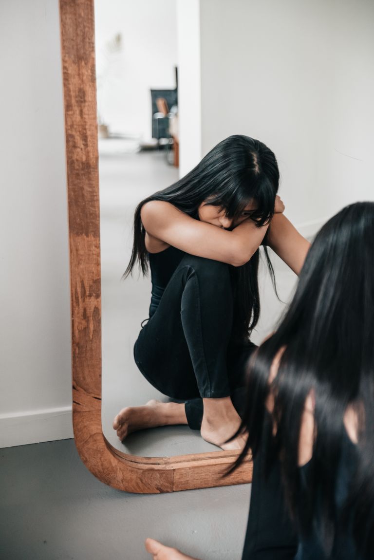A woman sitting pensively by a large wooden mirror indoors, reflecting on her thoughts.