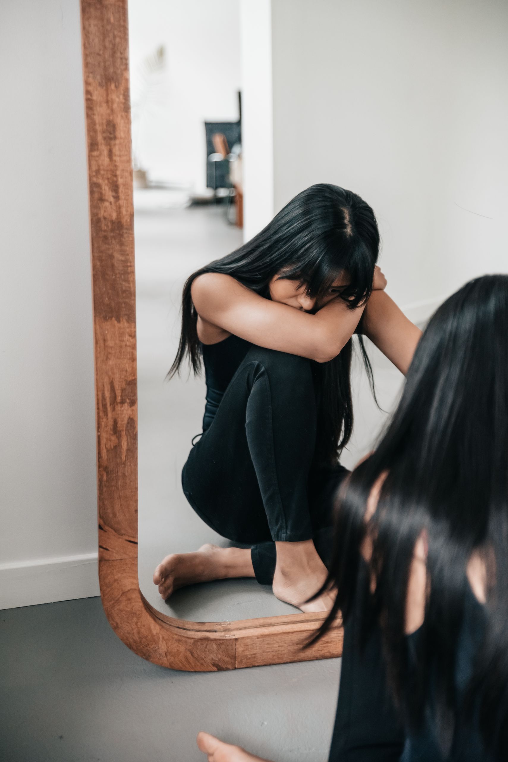 A woman sitting pensively by a large wooden mirror indoors, reflecting on her thoughts.