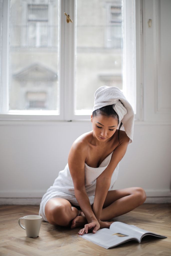 A woman wrapped in a towel reads by a window, epitomizing comfort and relaxation with a warm drink by her side.Self Love Rituals