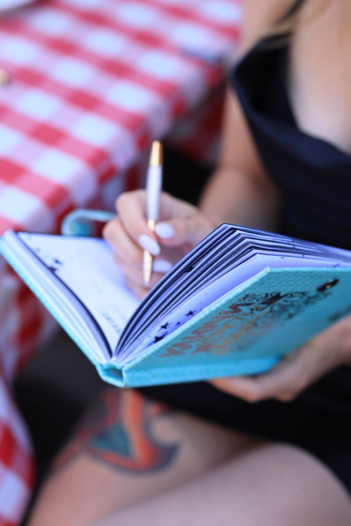 A woman writing in a blue notebook with a pen while sitting outdoors on a sunny day.