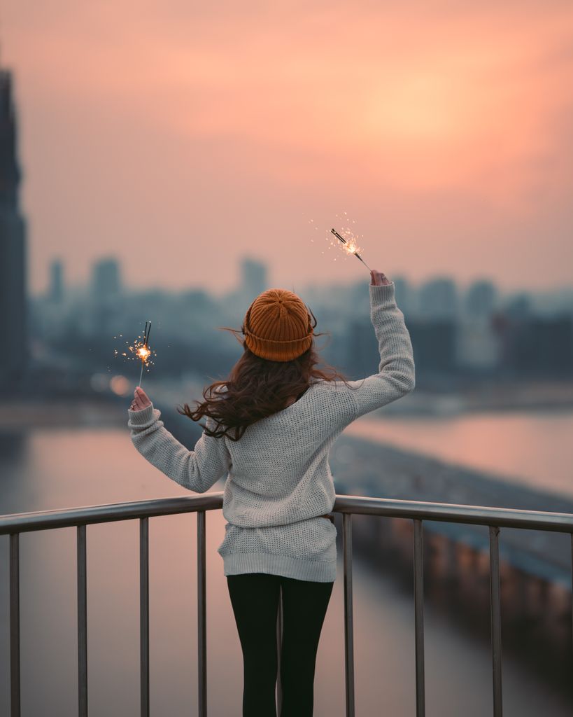 A young woman celebrates with sparklers on a balcony overlooking Seoul during a winter sunset.