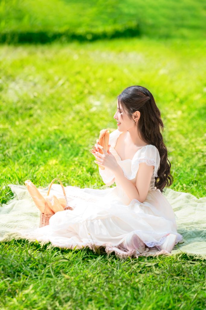 A young woman in a white dress enjoys a picnic on a sunny day in a lush green park.