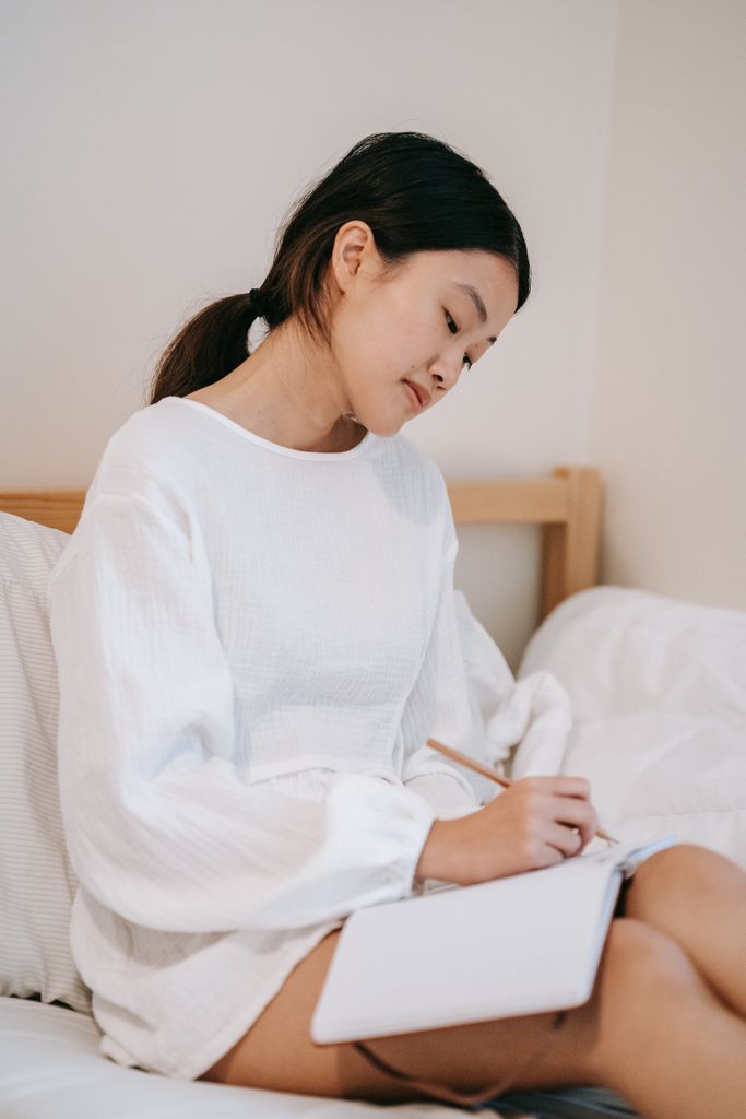 A young woman in a white sweater writing in a notebook while sitting on a bed.