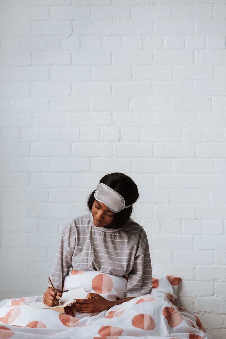 A young woman in striped pajamas and a sleep mask writes in a notebook while sitting on a bed with a polka-dot cover.
